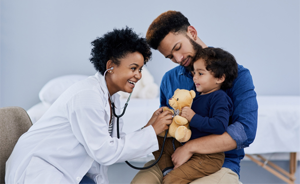 A dad holds his child, who is holding a teddy bear, while a doctor checks the bear's heart with a stethoscope.
